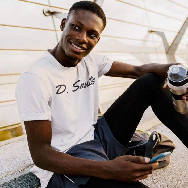 Man wearing a 'D. Snuts' t-shirt holding a water bottle and phone, sitting outdoors.